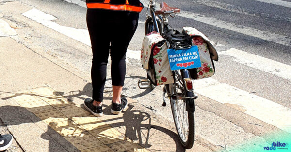 Ciclista mulher atravessa na faixa de pedestres. A placa na bicicleta lembra aos motoristas que sua filha a espera em casa.